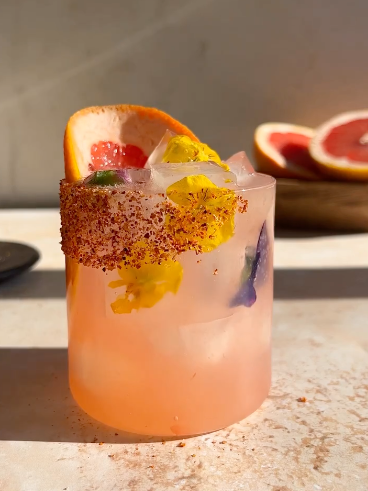 a light pink cocktail with edible flowers and a tajin rim
